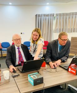 Photo of a young white woman pointing to a laptop screen an older white man is working on. Another white man is seated next to them using a computer.